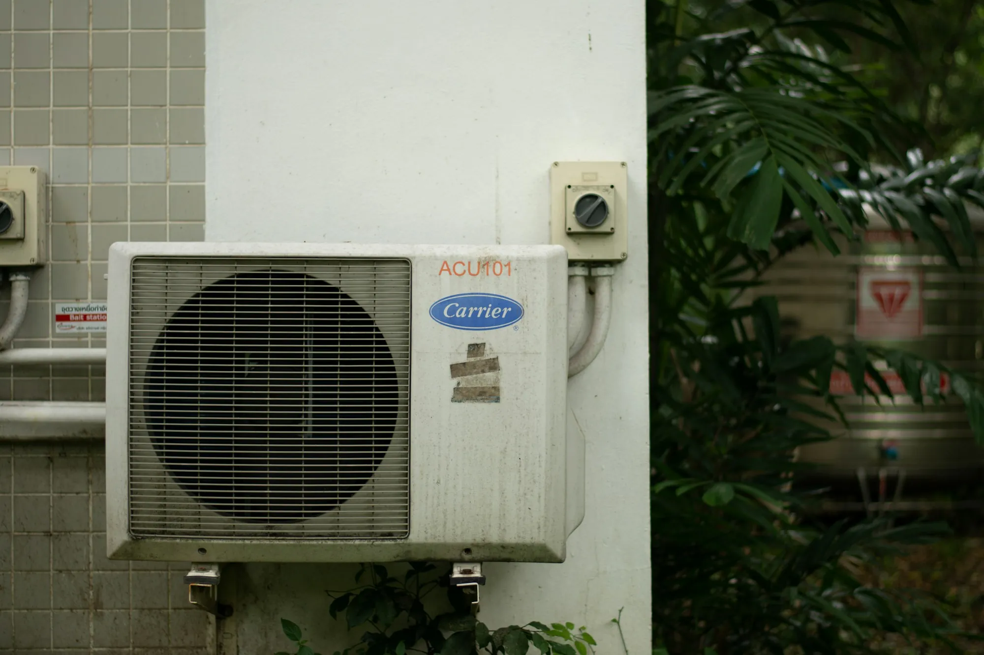 Outdoor air conditioner condenser on a wall beside green plants